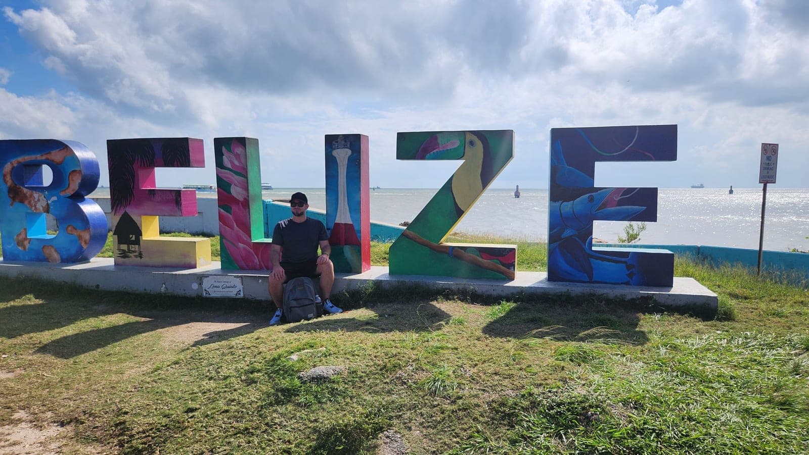 A visitor sitting in front of large colorful 'BELIZE' letters overlooking the water