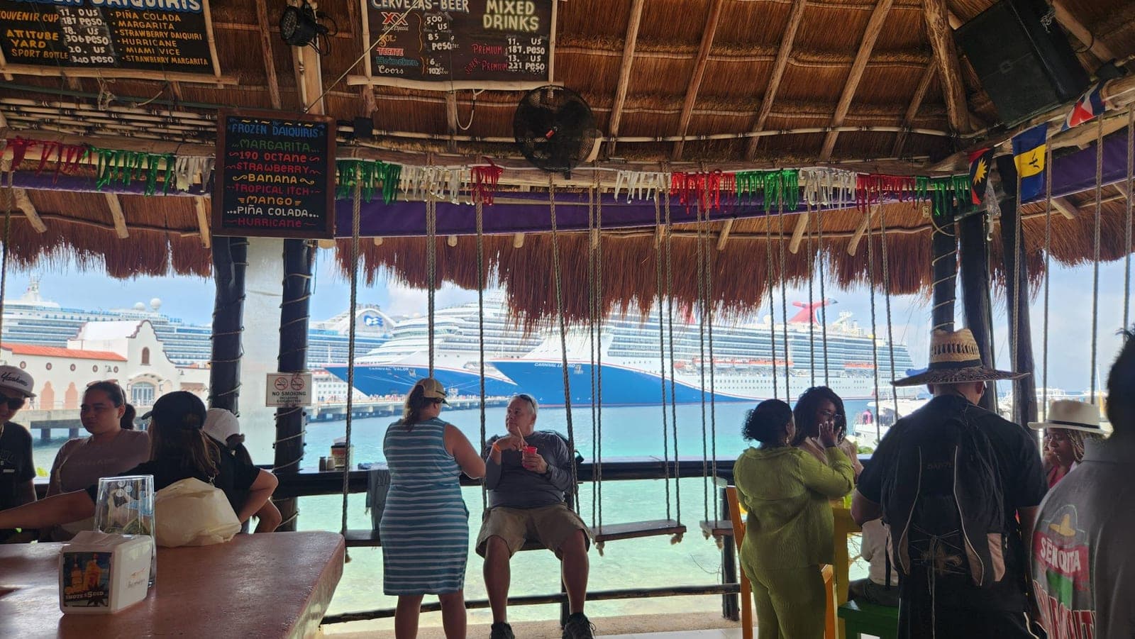 A thatched-roof beach bar with swing seats and a drink menu, looking out at cruise ships in Cozumel harbor