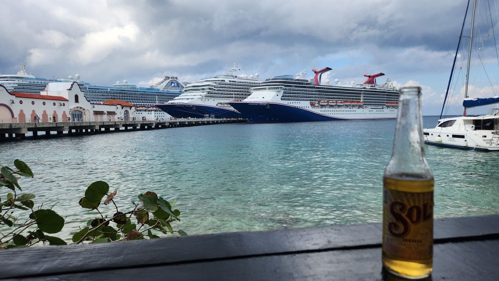 Two Carnival cruise ships docked in Cozumel harbor, viewed past a beer bottle on a waterfront table