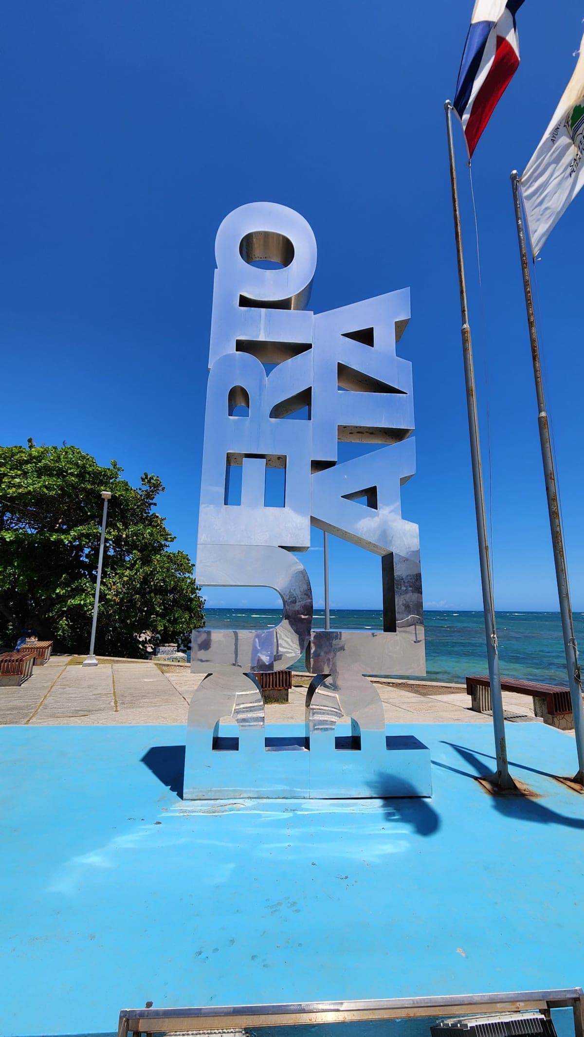 A tall metallic 'Puerto Plata' sign sculpture on the waterfront with the ocean in the background