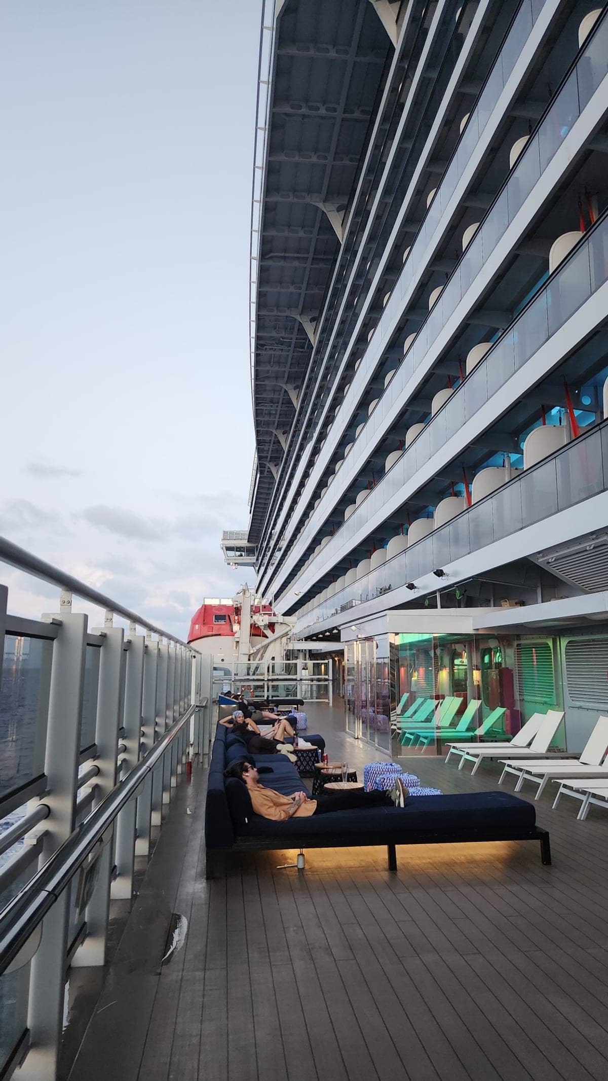 Guests relaxing on lounge daybeds along Valiant Lady's open deck at dusk, with a red lifeboat in view