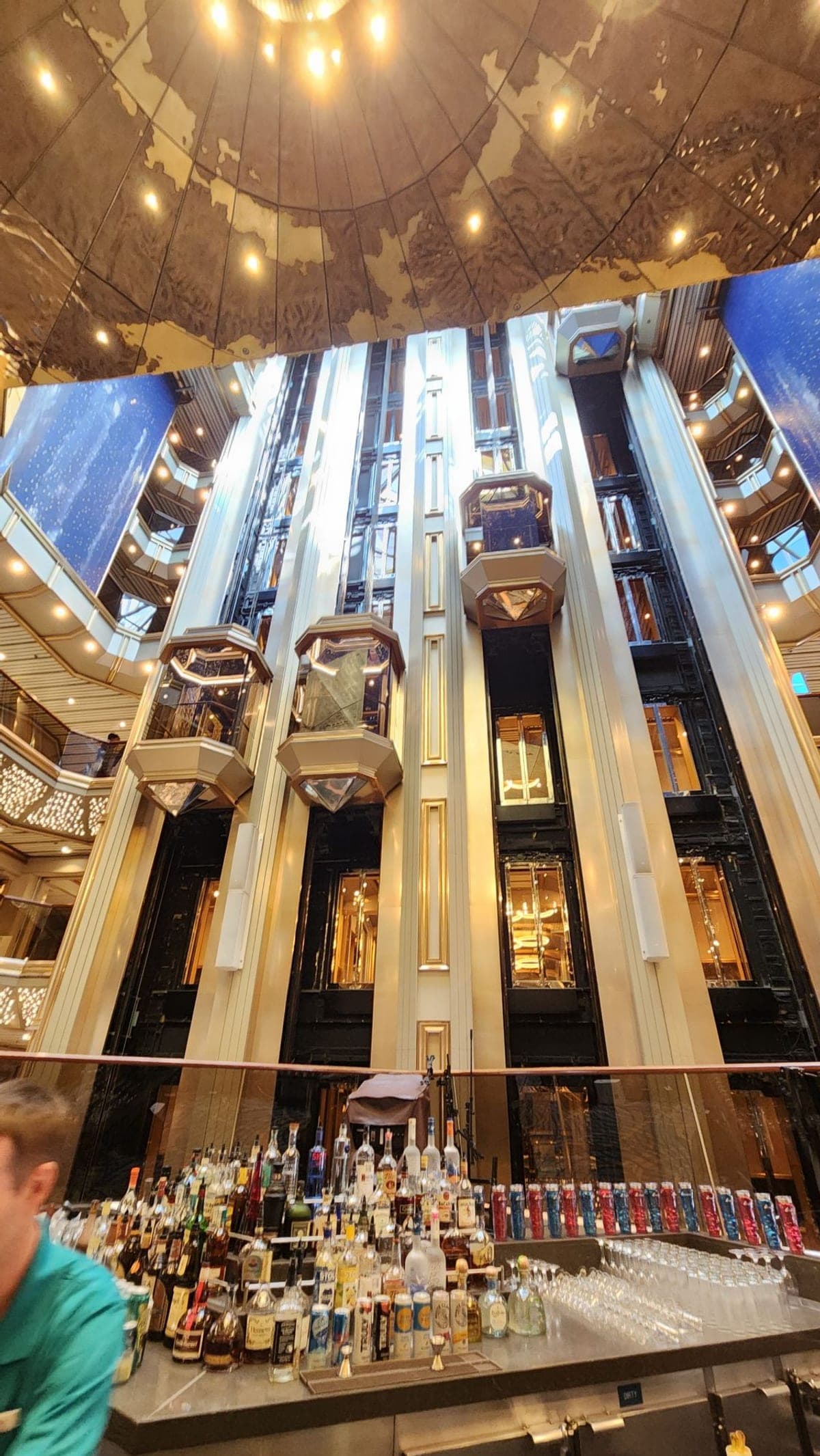 Looking up through Carnival Sunrise's tall atrium at glass elevators beneath a gold domed ceiling with a world map design