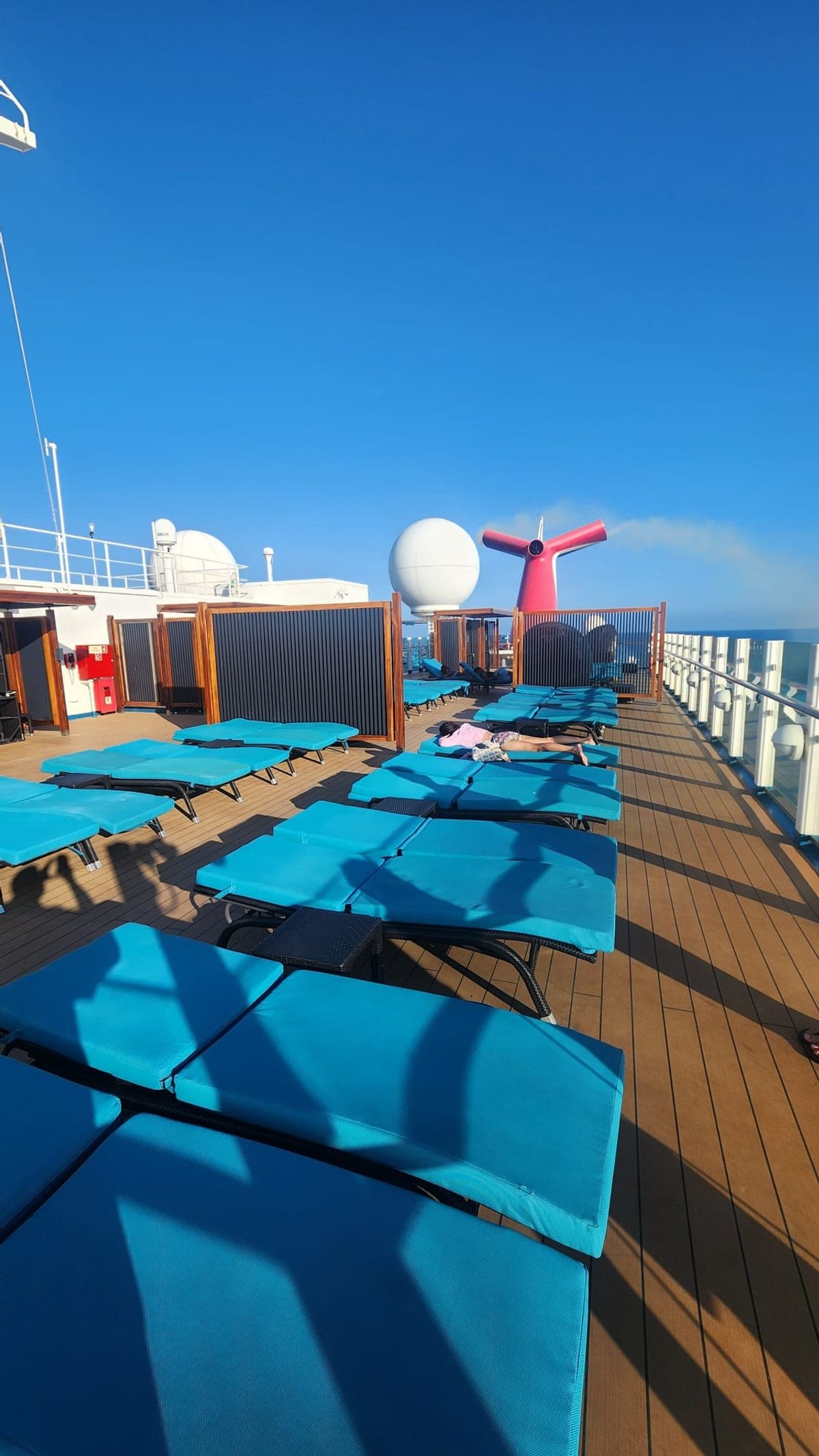 Rows of empty teal loungers on Carnival Sunrise's pool deck beside the ship's red funnel under a clear blue sky
