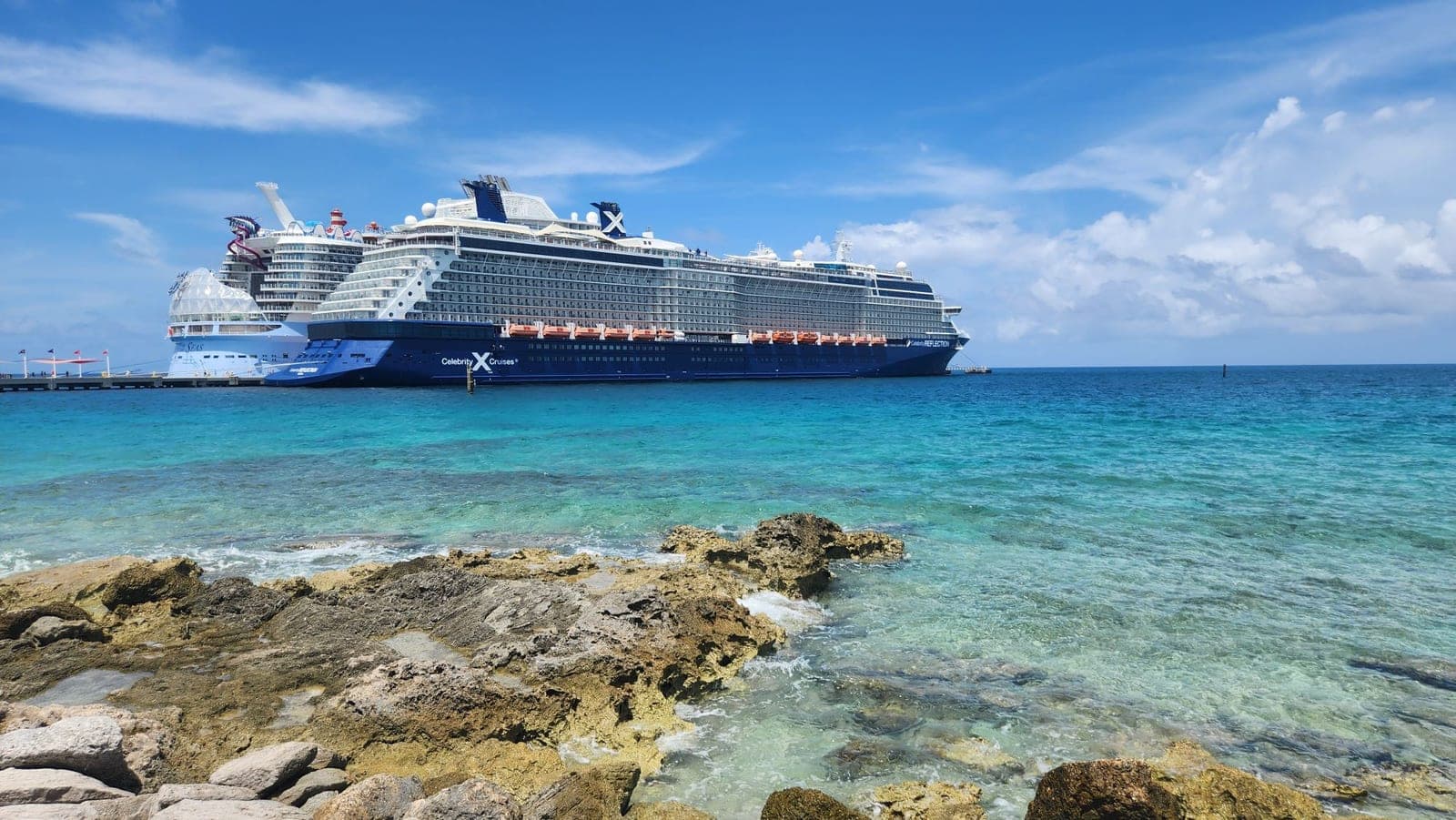 Celebrity Reflection anchored offshore near a rocky, turquoise-water coastline with another cruise ship nearby