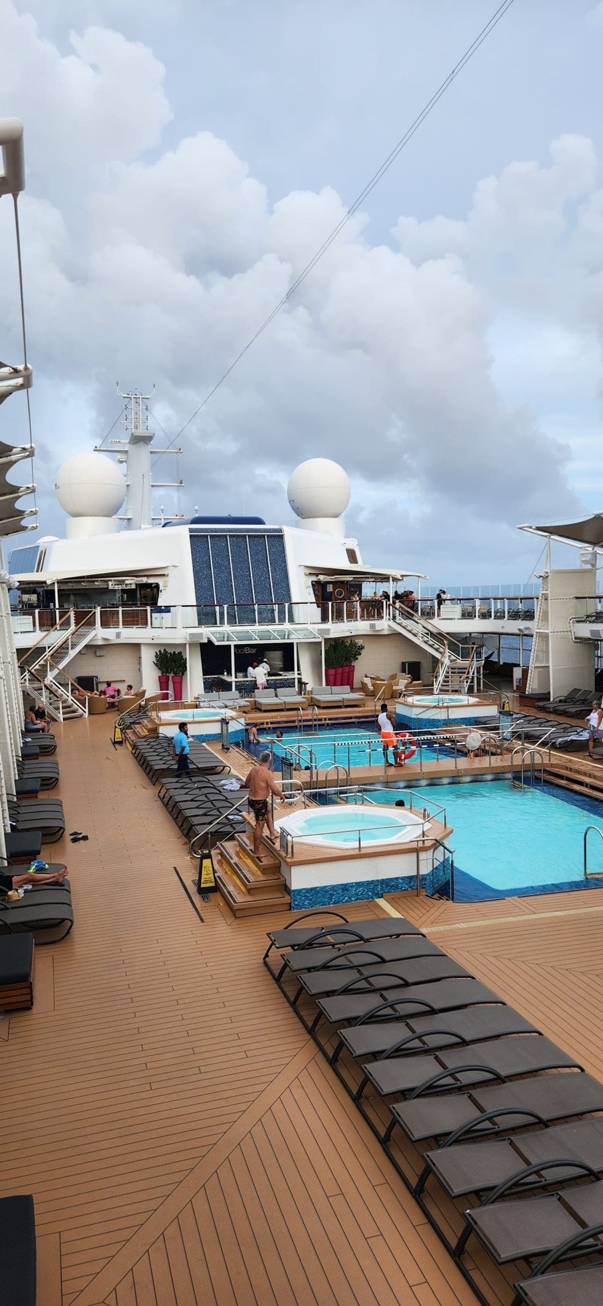 Celebrity Reflection's pool deck with two pools, hot tubs, and the pool bar under a cloudy sky