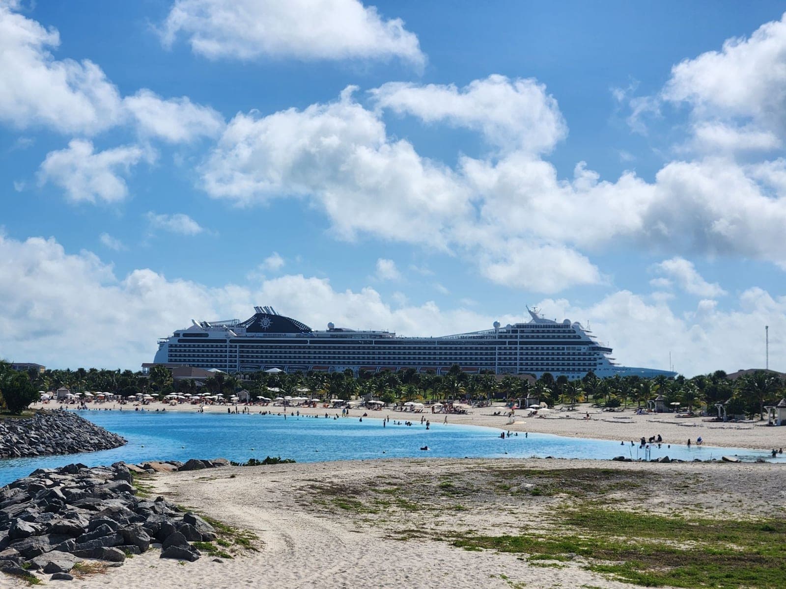 MSC Magnifica anchored offshore at a turquoise-lagoon beach with sunbeds and palm trees